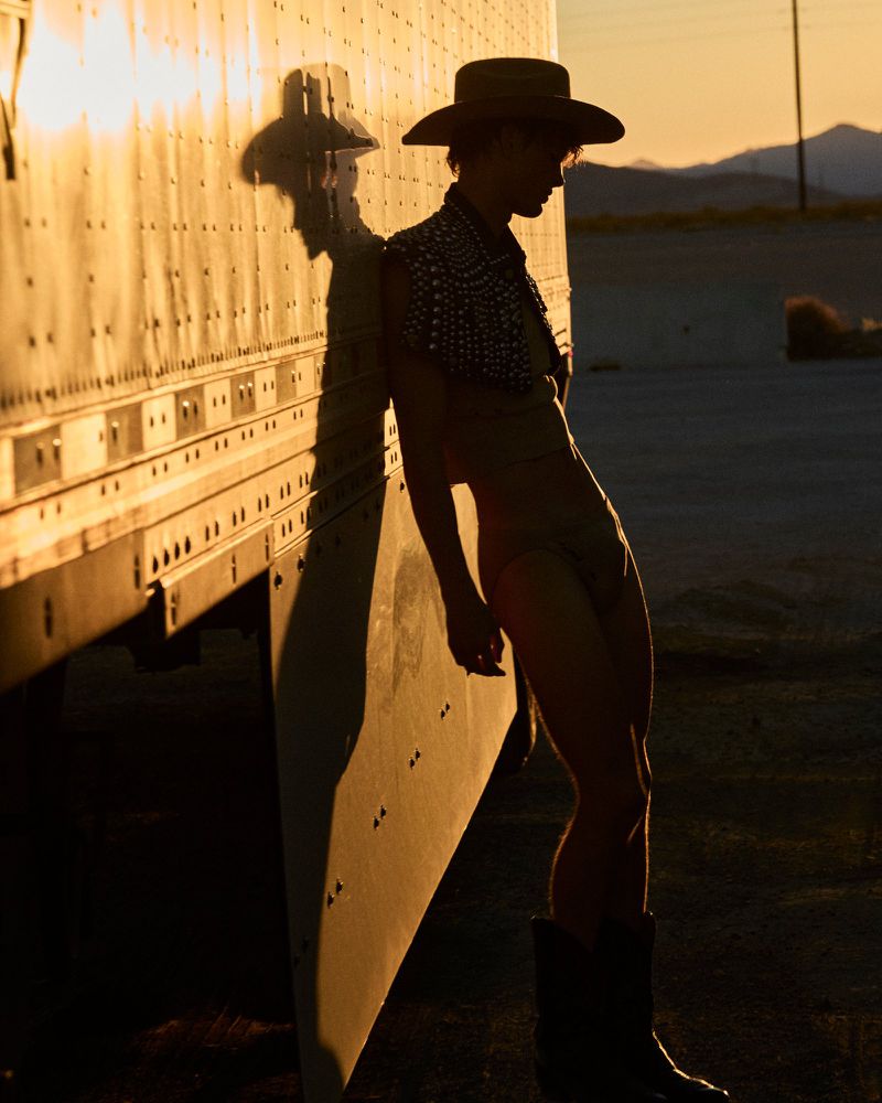 Man in hat and boots leans against semi-truck in warm light, with desert mountains in the background at golden hour.