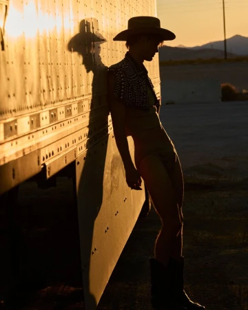 Man in hat and boots leans against semi-truck in warm light, with desert mountains in the background at golden hour.