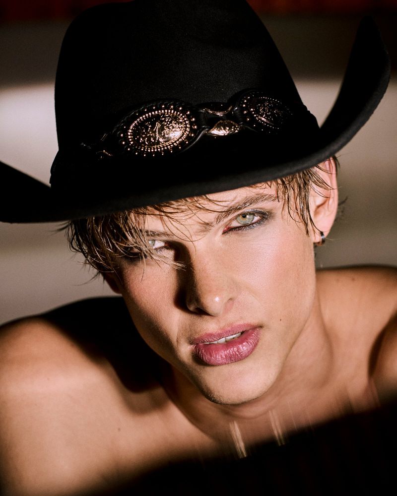 Portrait of a shirtless adult male with a black cowboy hat, smokey eye makeup, light colored hair, and light skin tone in studio lighting.