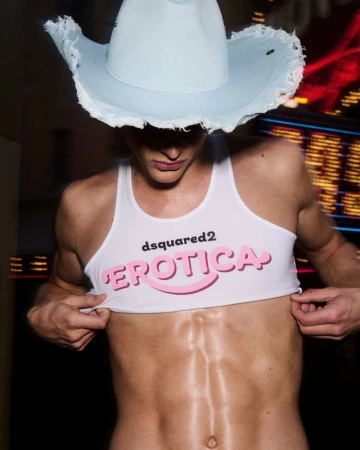 Male fashion model wearing a denim cowboy hat and a crop top with 'Erotica' written on it, standing indoors with blurred background lights.