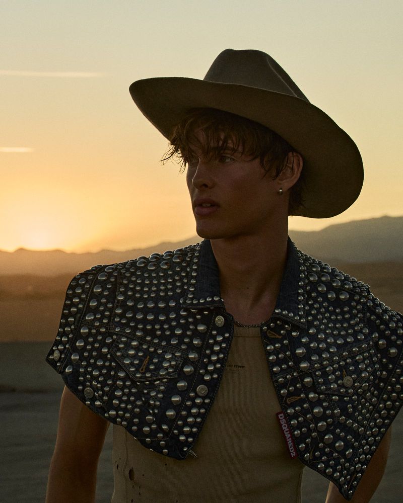 Portrait of a handsome man in a cowboy hat and studded denim vest, standing outdoors against a sunset with mountains in the distance.