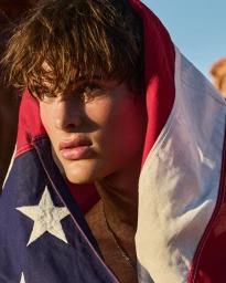 Young male model wrapped in an American flag with direct sunlight illuminating his face against a natural outdoor setting