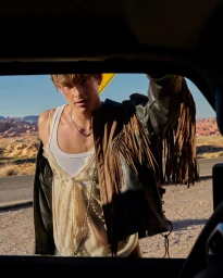 Young man in a fringed leather jacket and sheer top leaning through a car window in a sunny desert landscape.