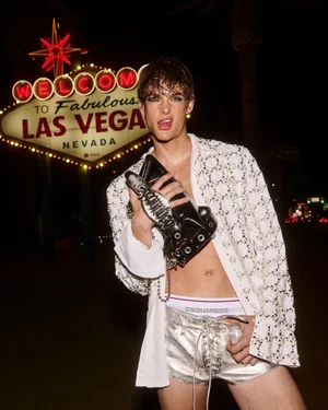 A male model posing in an open white shirt and metallic silver shorts in front of the Welcome to Fabulous Las Vegas sign at night.