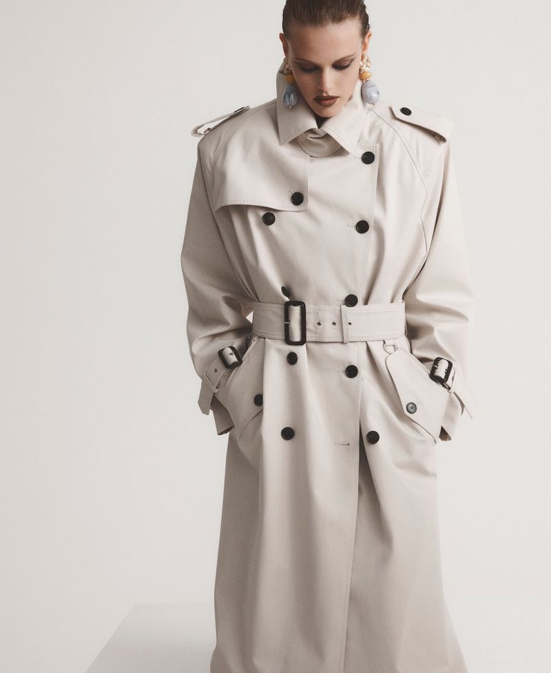 Model wearing an oversized cream double-breasted trench coat with a buckled belt against a white studio background.