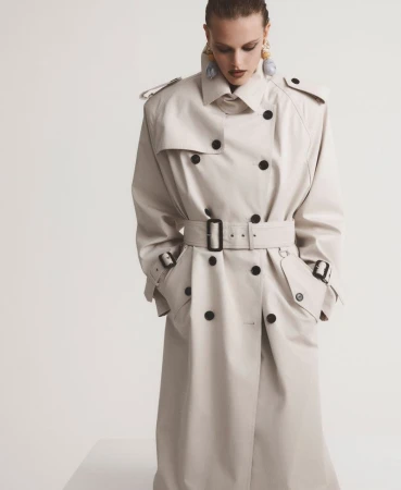 Model wearing an oversized cream double-breasted trench coat with a buckled belt against a white studio background.