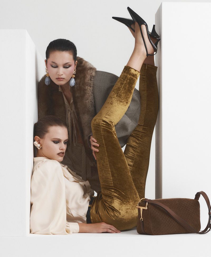 Two fashion models in a white corner studio setting wearing elegant vintage clothing, one with her legs raised, and a brown purse.