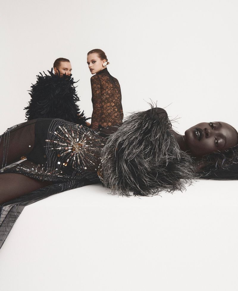 Three women pose in high fashion, showcasing black embellished dresses and feather accents against a minimalist white backdrop.