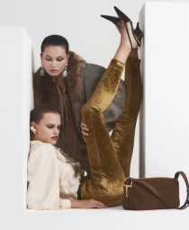 Two models pose between white geometric blocks, one reclining in gold velvet pants and the other wearing a fur-trimmed jacket.