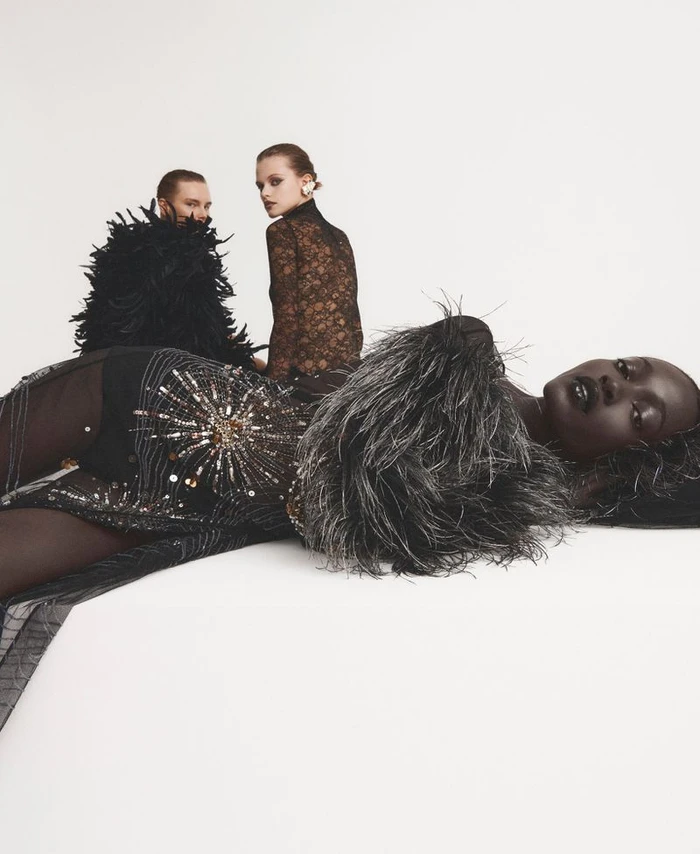 A Black model in an embellished black feather dress lying down on a white seamless backdrop with two models posing behind her.