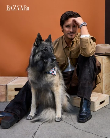 Diego Boneta sits beside his dark-haired dog wearing a patterned bandana; he wears a khaki shirt and black boots in a studio shot.