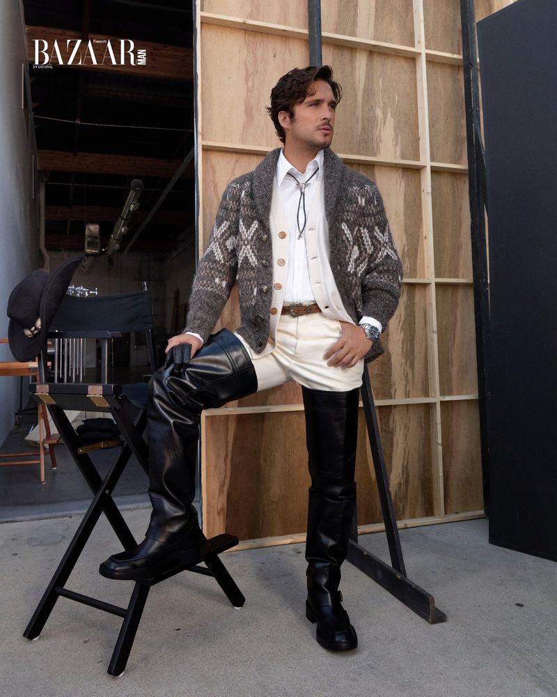 Man in patterned cardigan, cowboy bolo tie, and tall black boots poses next to a director's chair in a photo studio setting.