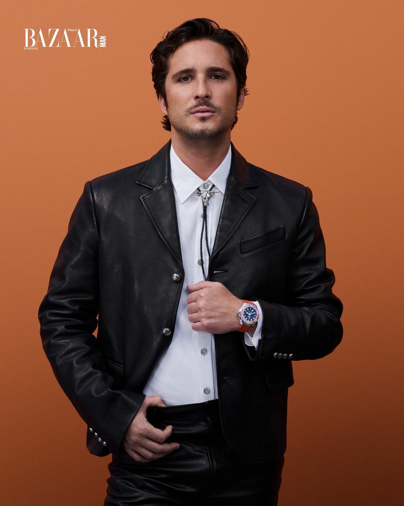 Portrait of a man in a black leather jacket, white shirt, and bolo tie against a warm orange studio backdrop for Bazaar.