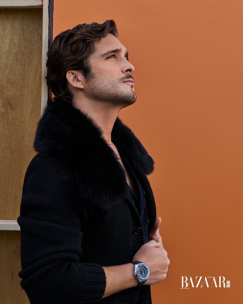 Man in dark jacket with fur collar looks upward against an orange wall, showcasing a wristwatch and subtle side profile.