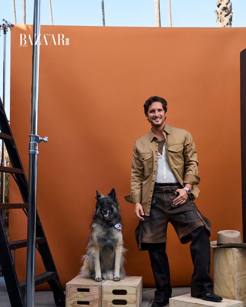 Diego Boneta poses in a studio with his dog in front of an orange backdrop, with palms visible in the background and a Bazaar logo.
