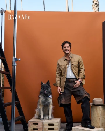 Diego Boneta poses in a studio with his dog in front of an orange backdrop, with palms visible in the background and a Bazaar logo.