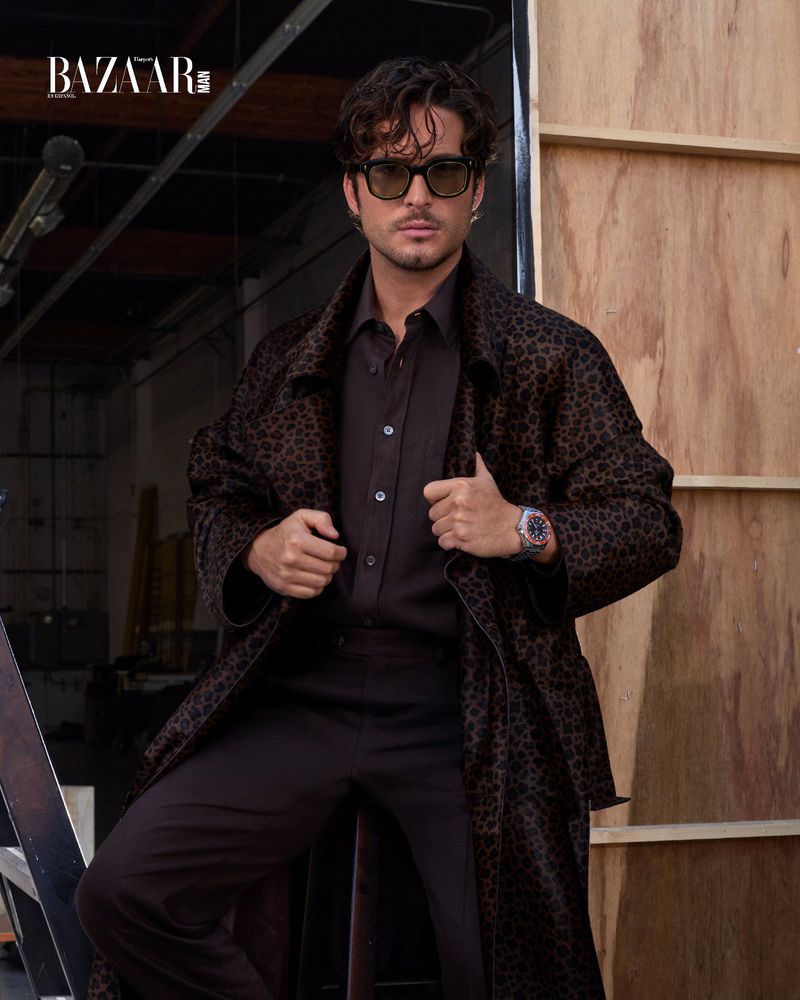 An adult male in a brown patterned coat and sunglasses poses in front of a wooden wall, featuring the Bazaar logo.