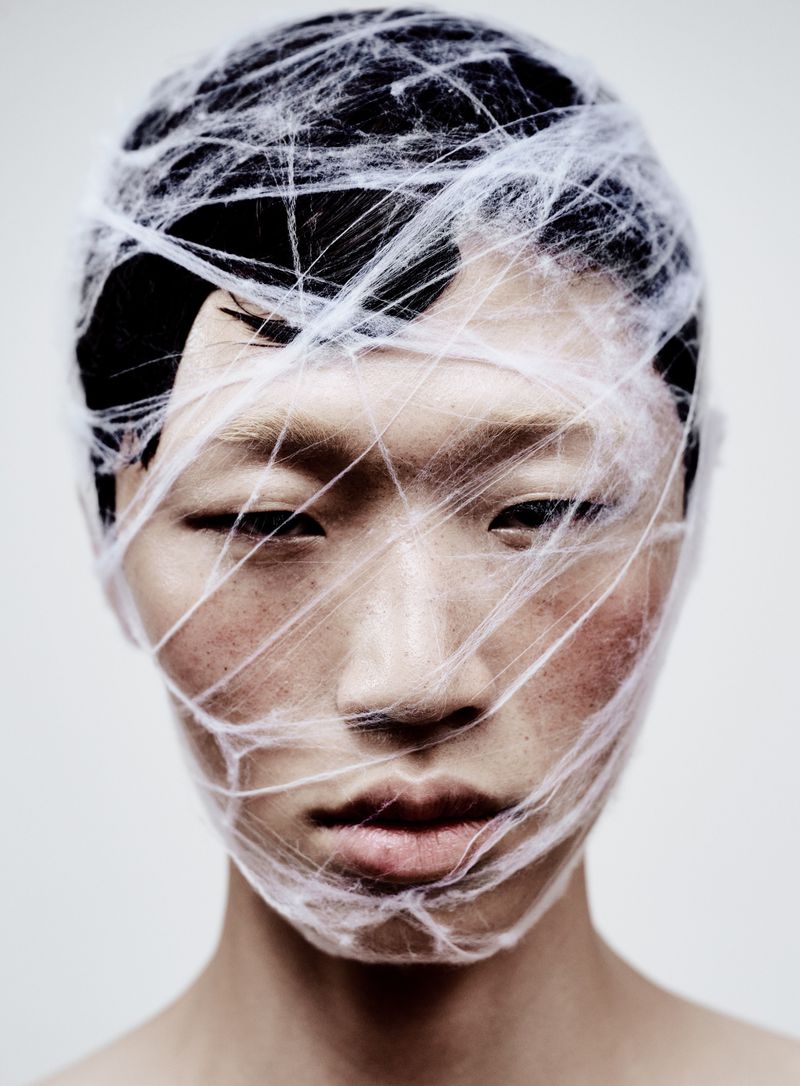 Striking headshot of an Asian model with dark hair, face closely wrapped in white threads, captured in a clean, brightly lit studio setting.