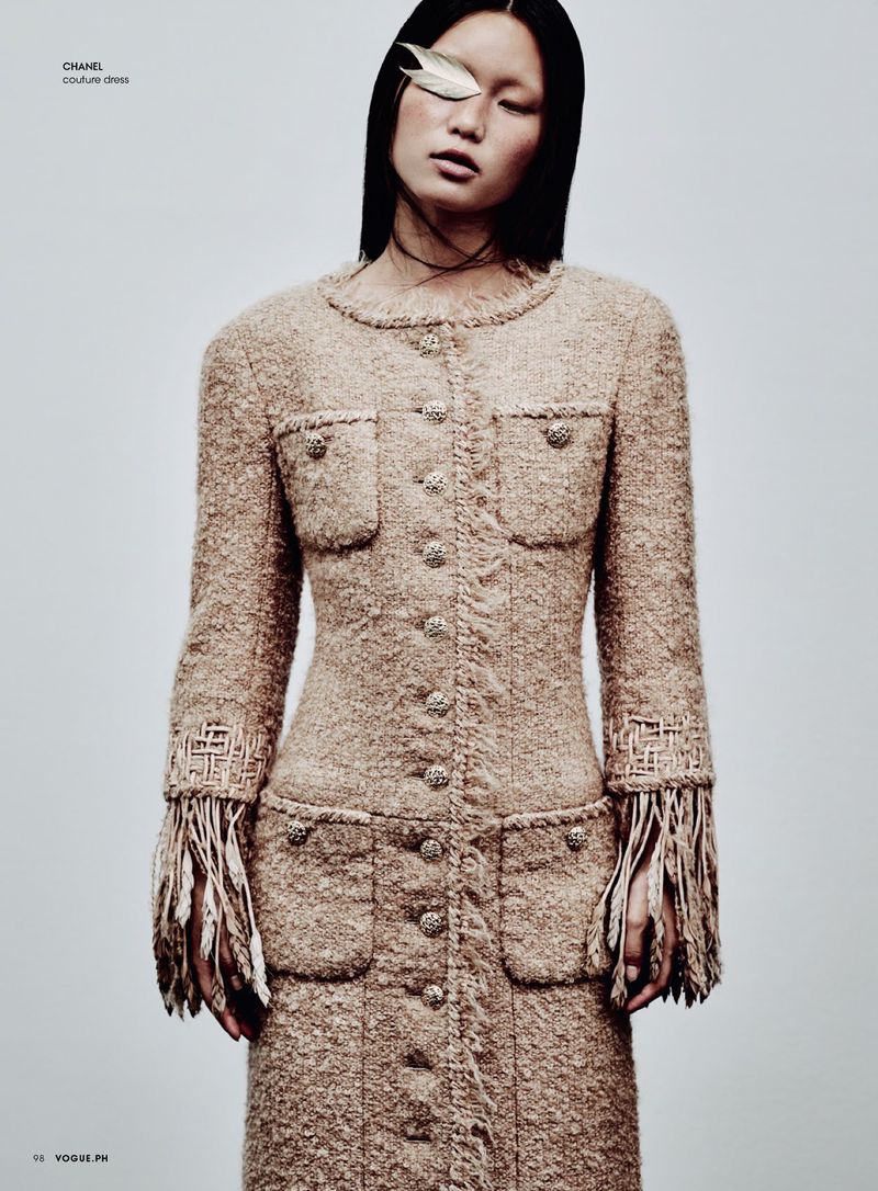 Fashion editorial shot of an Asian woman with a feather over her eye wearing a tweed coat dress with fringe against a light background.