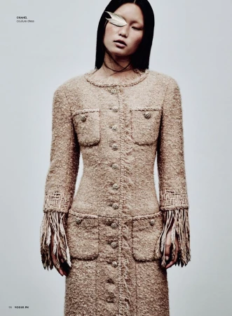 Fashion editorial shot of an Asian woman with a feather over her eye wearing a tweed coat dress with fringe against a light background.