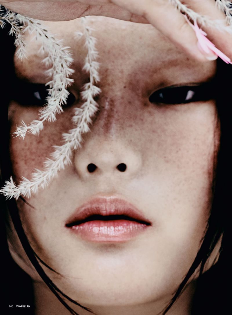 Dramatic close-up portrait of an Asian woman with freckles and black hair, accentuated by white floral accents against a dark backdrop.