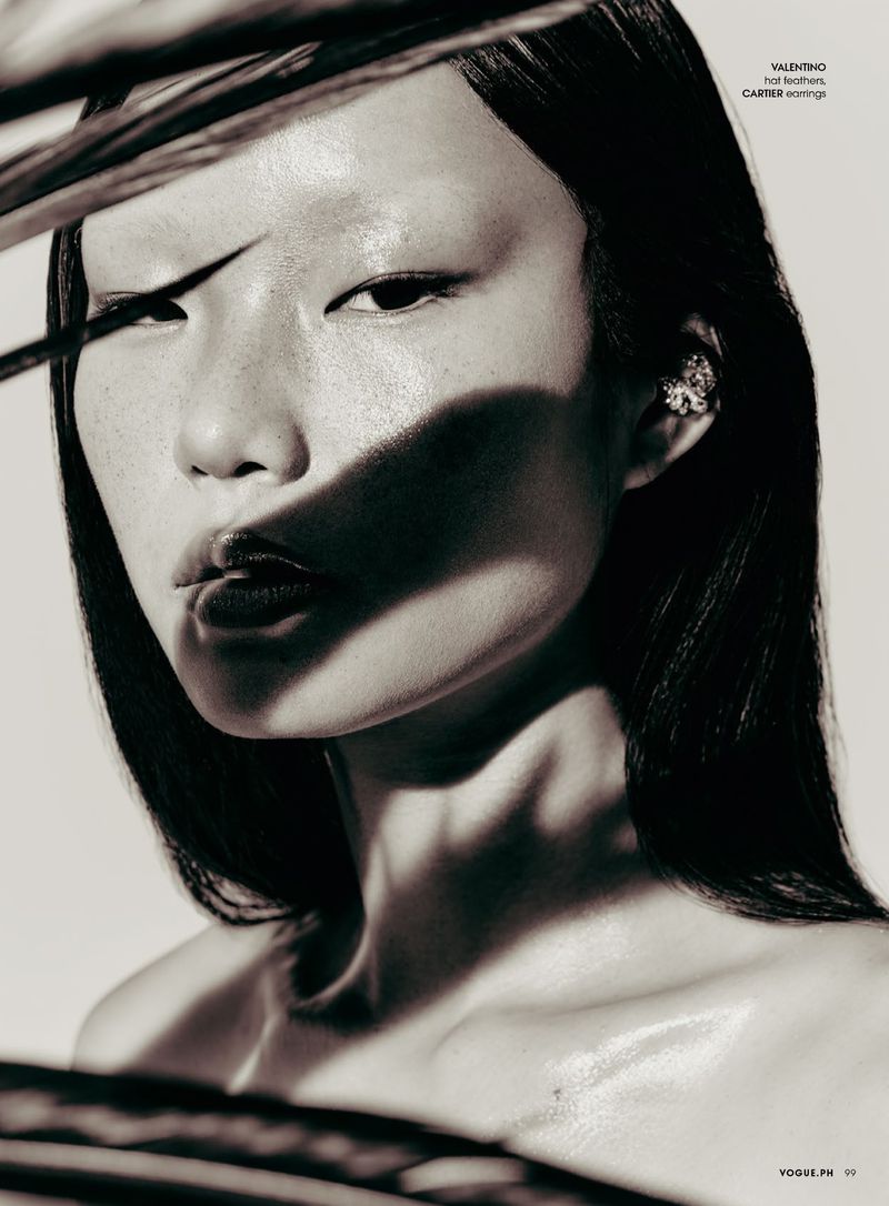 Close-up portrait of an Asian woman with sleek black hair and shiny lips, adorned with elegant earrings and accented by shadows and leaves.