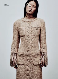 Model with long dark hair wearing a textured beige tweed coat against a plain light grey studio background.