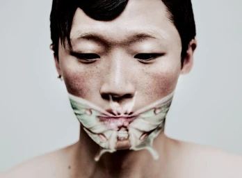 Close-up portrait of an East Asian male model looking down with a pale green moth pattern across his lower face and mouth.