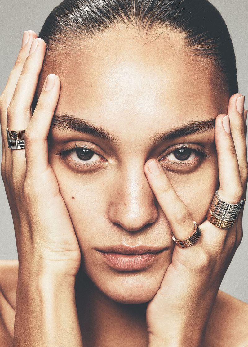 Close-up portrait of a young adult woman with tan skin, styled hair, and multiple gold and silver rings, posing in a studio setting.