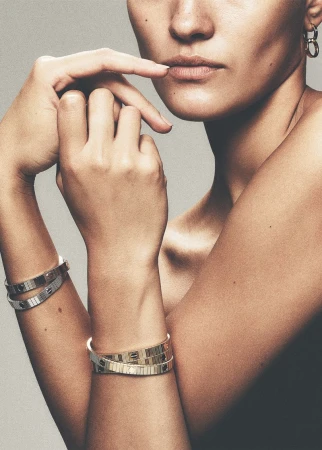 Close-up of model Aida Atarssa showcasing layered metallic bangles on both wrists with a neutral studio backdrop.