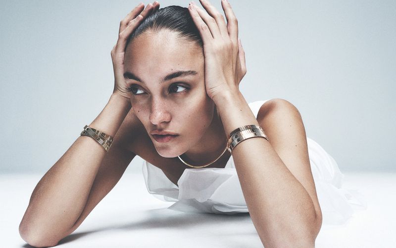 Portrait of a woman lying down wearing a white dress and gold bracelets against a gradient gray background in a studio.