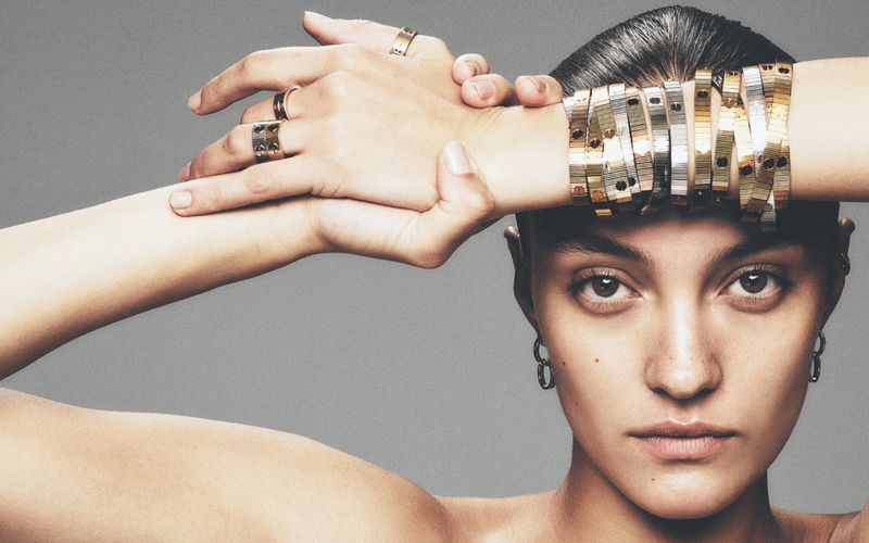 Close-up studio shot of a woman with short hair and jewelry including rings and bracelets against a solid grey backdrop in portrait orientation.