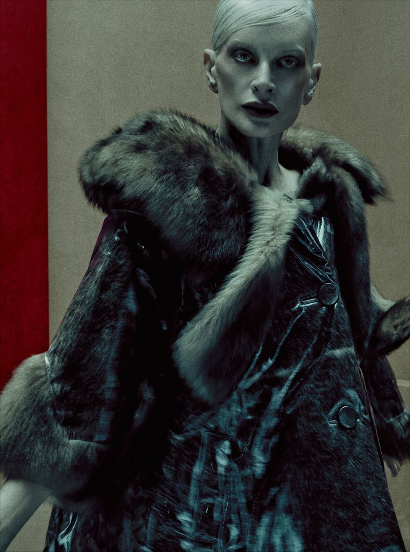 Fashionable woman in fur-trimmed coat, studio setting, neutral backdrop with red accent. Dark makeup, short hair, moody atmosphere.