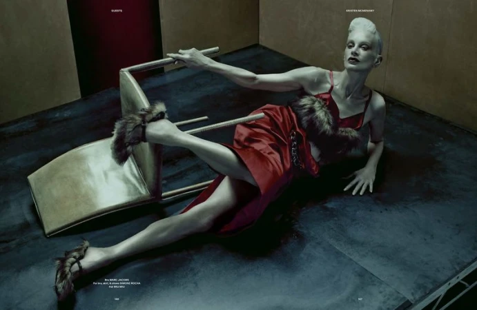 Fashion shot of a reclining adult woman in a red dress and fur-lined sandals with a fallen chair in an indoor, studio-like environment.