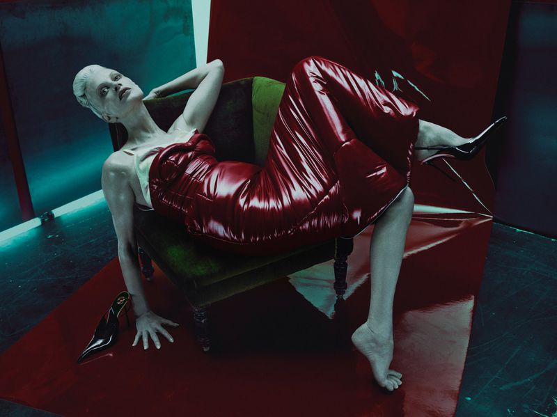 A woman in a red dress reclines on a green chair in a staged studio environment with glossy red and turquoise backgrounds.
