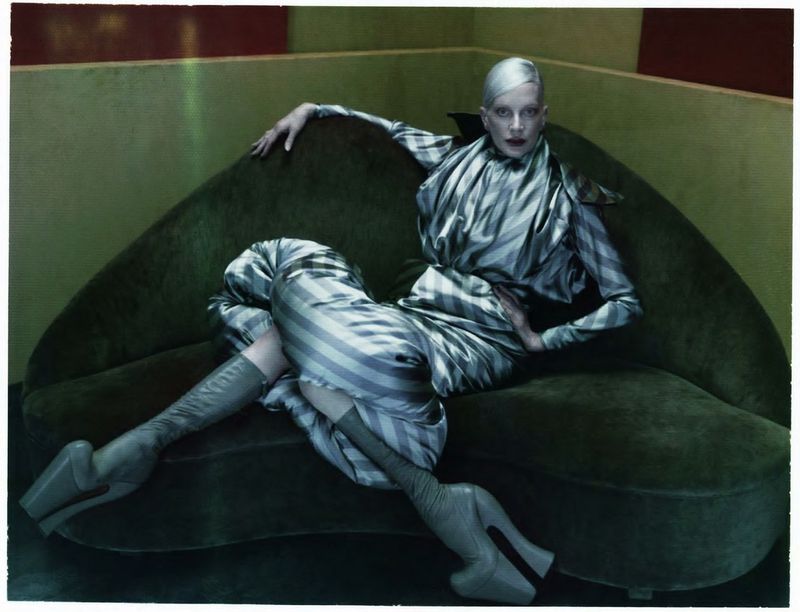 A fair-skinned adult female model lounges on a green couch, wearing a silver-striped dress and matching boots in a studio setting.