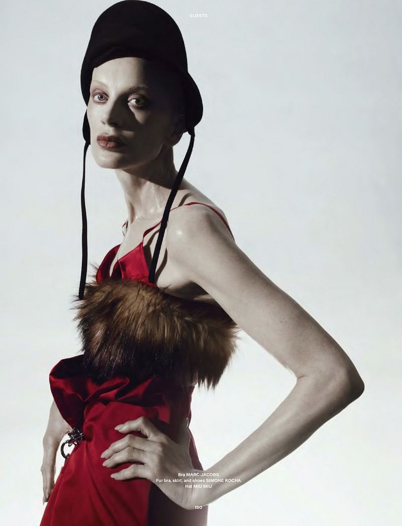 A fashion portrait features a slender adult female model in a red dress with fur trim and a black hat posing against a bright background.