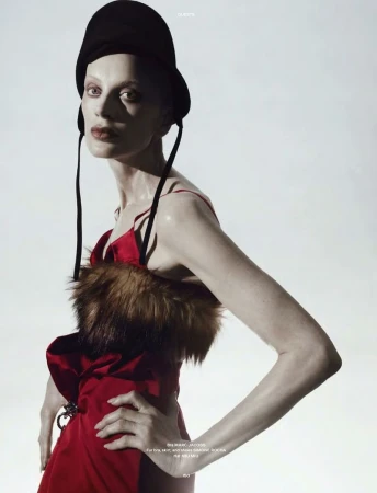 A fashion portrait features a slender adult female model in a red dress with fur trim and a black hat posing against a bright background.
