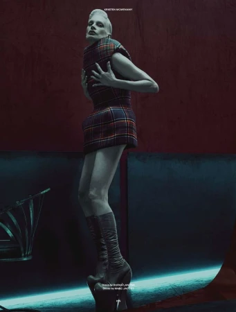 Woman in plaid dress and platform boots in a studio, striking an unusual pose with hands near chest, editorial fashion photography.