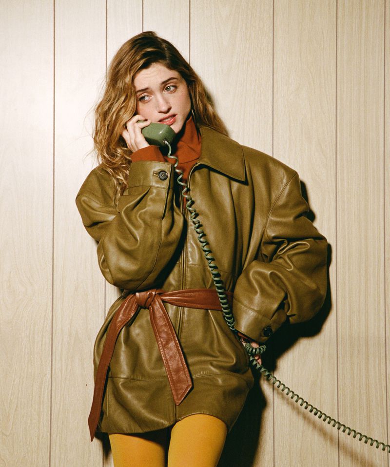 A woman with wavy blonde hair is talking on a green vintage telephone wearing a brown leather jacket with a sash in a studio.