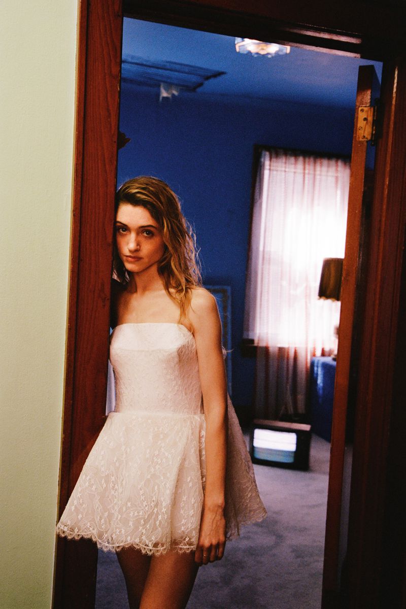 Natalia Dyer standing in a wooden doorway wearing a white strapless lace mini dress inside a blue-walled interior.