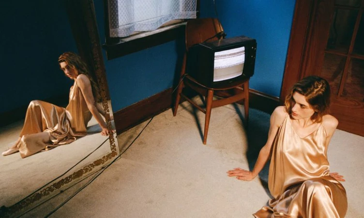 A young woman in a gold satin dress sits on the floor next to a floor-length mirror in an indoor setting with a vintage television on a chair.