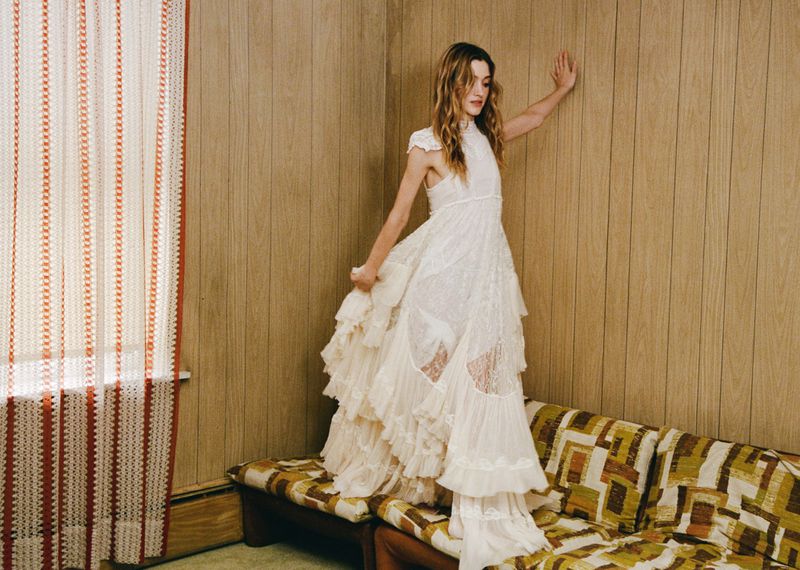 A woman poses in a white lace dress with a tiered skirt indoors near paneled walls and a patterned couch, creating a vintage editorial fashion shot.