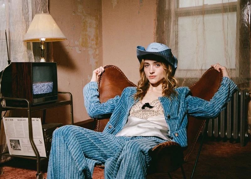 Stylish woman in blue corduroy suit and hat, seated in vintage armchair near a retro TV set in an indoor setting with warm lighting.