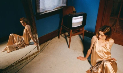 Natalia Dyer seated on a carpeted floor wearing a gold satin slip dress beside a vintage television in an editorial setting.
