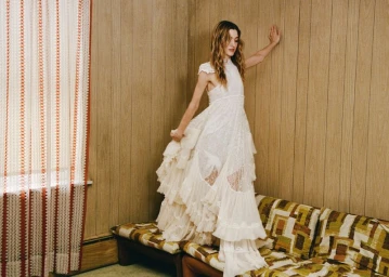 Natalia Dyer standing on a retro patterned sofa in an off-white tiered lace gown against a wood-paneled wall.