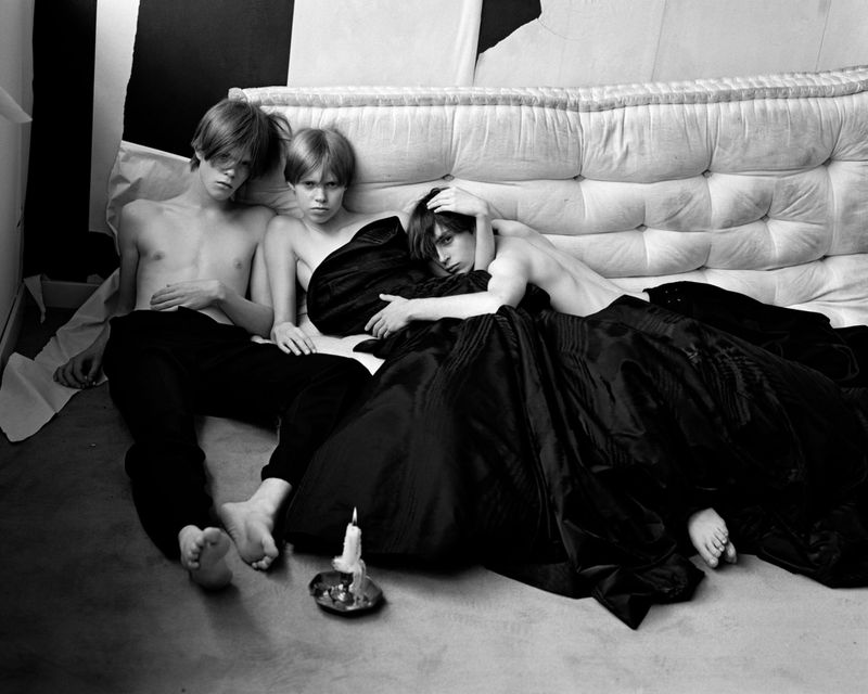 Three young adults pose indoors on a cushioned bed under a black satin sheet with a lit candle in a black and white photograph.