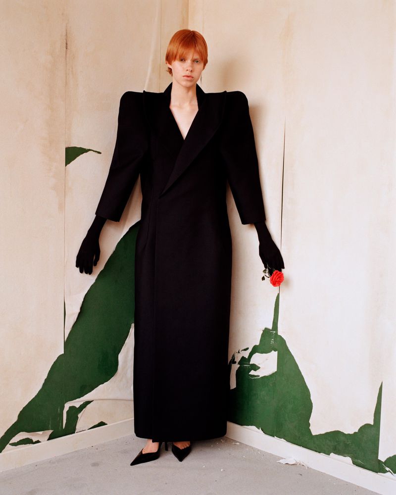 A model poses in a long black dress and gloves, holding a red rose, against a backdrop of cream and green painted walls in a studio setting.