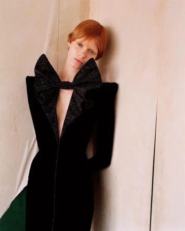 An adult female poses indoors wearing a black velvet couture gown with a large decorative bow detail in a fashion studio setting.