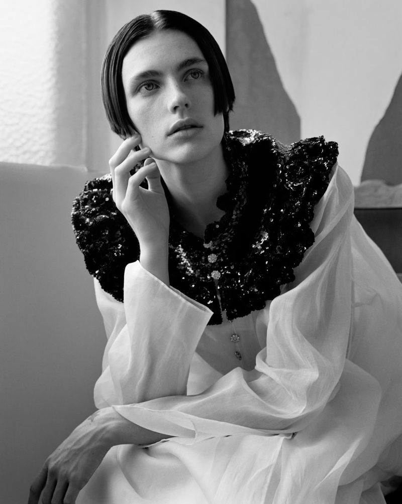 Monochrome portrait of a young adult with dark hair in a white top and black sequined collar, posing indoors with natural lighting.
